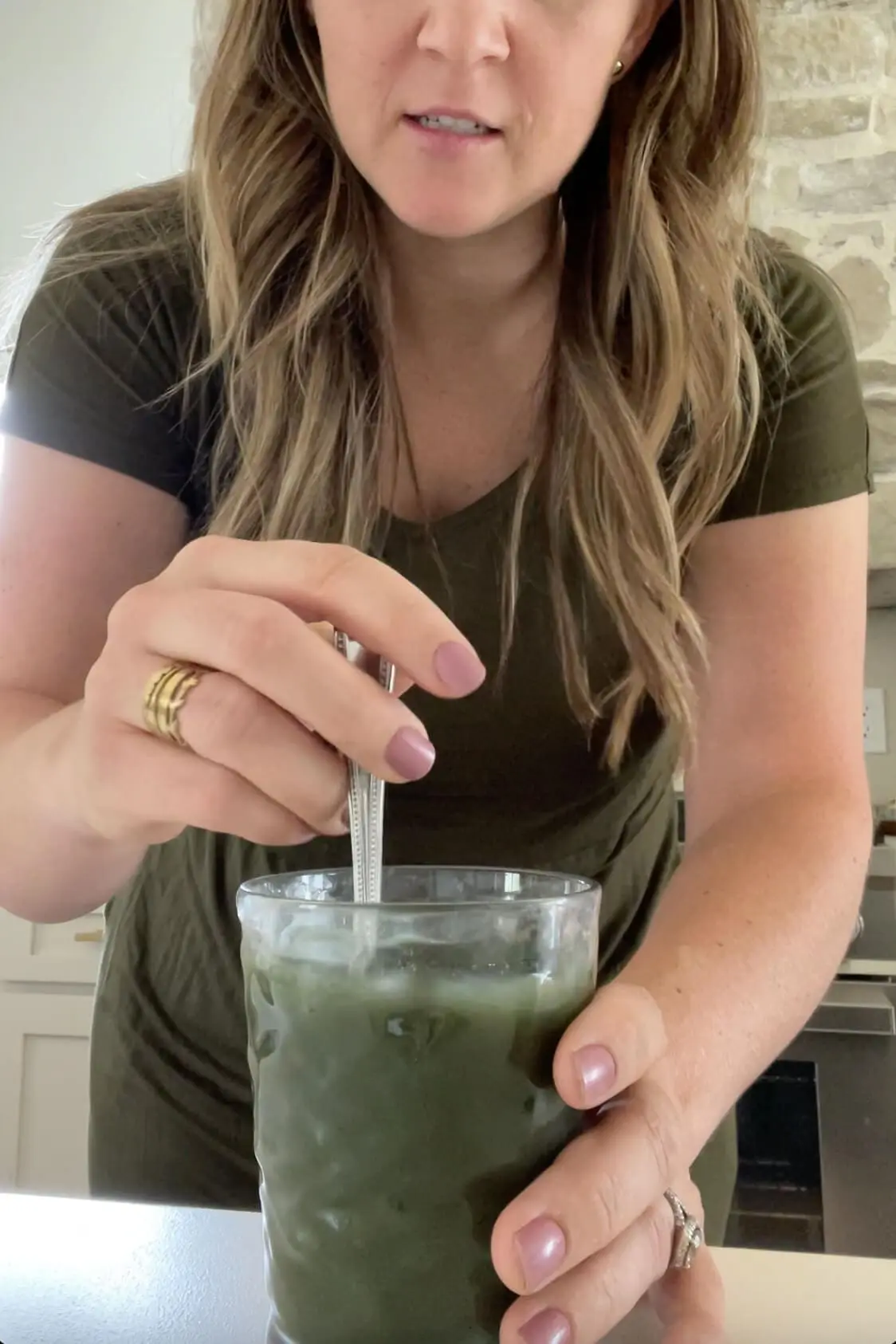 Close-up of a woman stirring a glass of iced AG1 greens powder, showing her wellness routine in a modern kitchen.